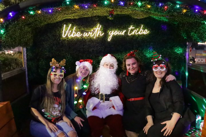 Four people with Santa, wearing festive attire, under a sign saying 'Vibe with your tribe' with colorful lights.