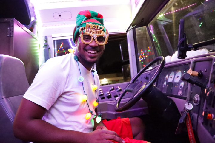 Man in festive hat and Merry Christmas glasses, sitting in a decorated vehicle, smiling.
