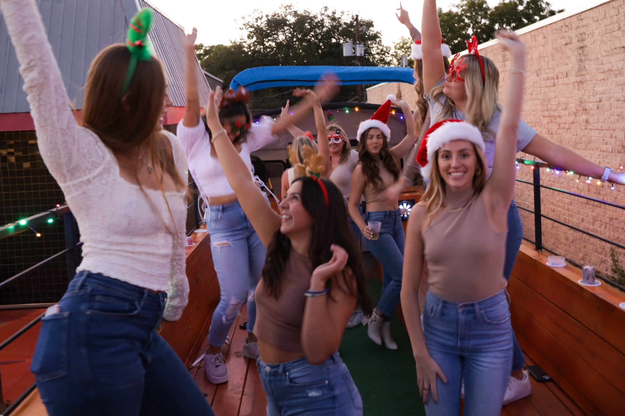 Group of people dancing in festive attire with holiday headbands on a decorated vehicle.