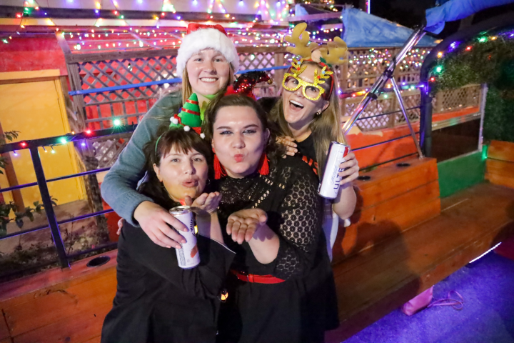 Four people in festive attire pose with drinks, surrounded by colorful lights.