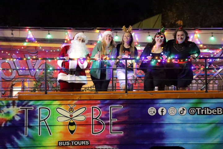 People in holiday costumes on a bus decorated with colorful lights and a 'Tribe Bus Tours' sign.