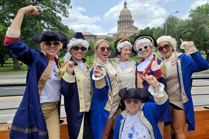Seven people in historical costumes pose cheerfully in front of a domed building on a sunny day.