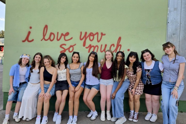 Group of twelve people smiling in front of a wall with 'I love you so much' graffiti.